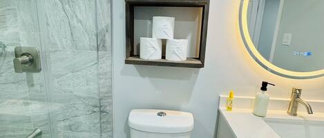 A bathroom with a glass shower enclosure, a toilet, a sink with a mirror, and a shelf with toilet paper.