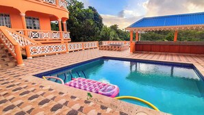 A swimming pool with a tiled deck, a covered seating area, and a building with orange columns and a blue roof. - Union Hilltop Paradise Villas - Premium Poolside Villa with Scenic Views (Castries)
