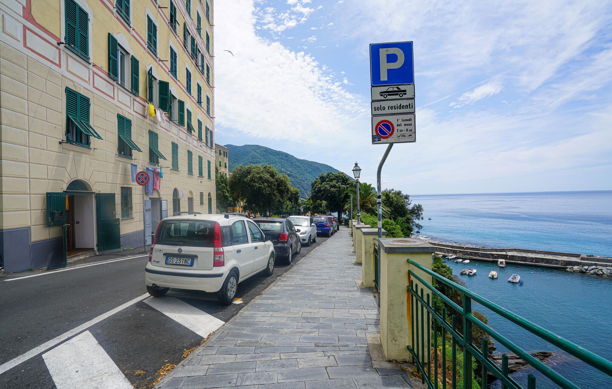 Eine Küstenstraße mit parkenden Autos, einem blauen Parkschild und Blick auf das Meer.
