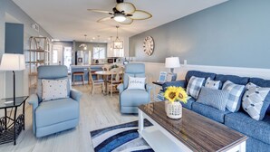 A modern living room with a blue sofa, two armchairs, a coffee table, and a dining area with a chandelier. - Run Away Bay 181 by SeaBreeze Vacation (Bradenton Beach)