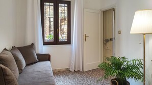 Een kamer met een bank, een raam met gordijnen, een deur, een lamp en een plant.