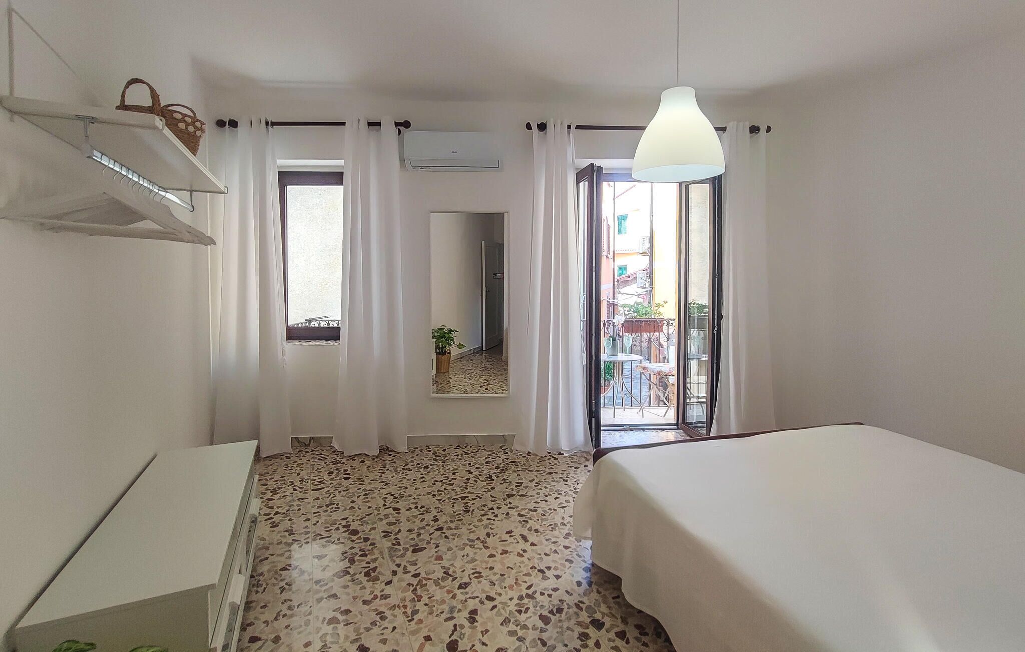A hotel room with a bed, a small table, a hanging basket, a window with curtains, and a view of the outside balcony.