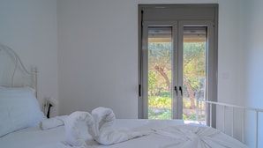 Ein ordentlich bezogenes Bett mit weißen Leinen und Handtüchern, ein weißer Kopfteil und Blick ins Grüne durch eine Glastür.