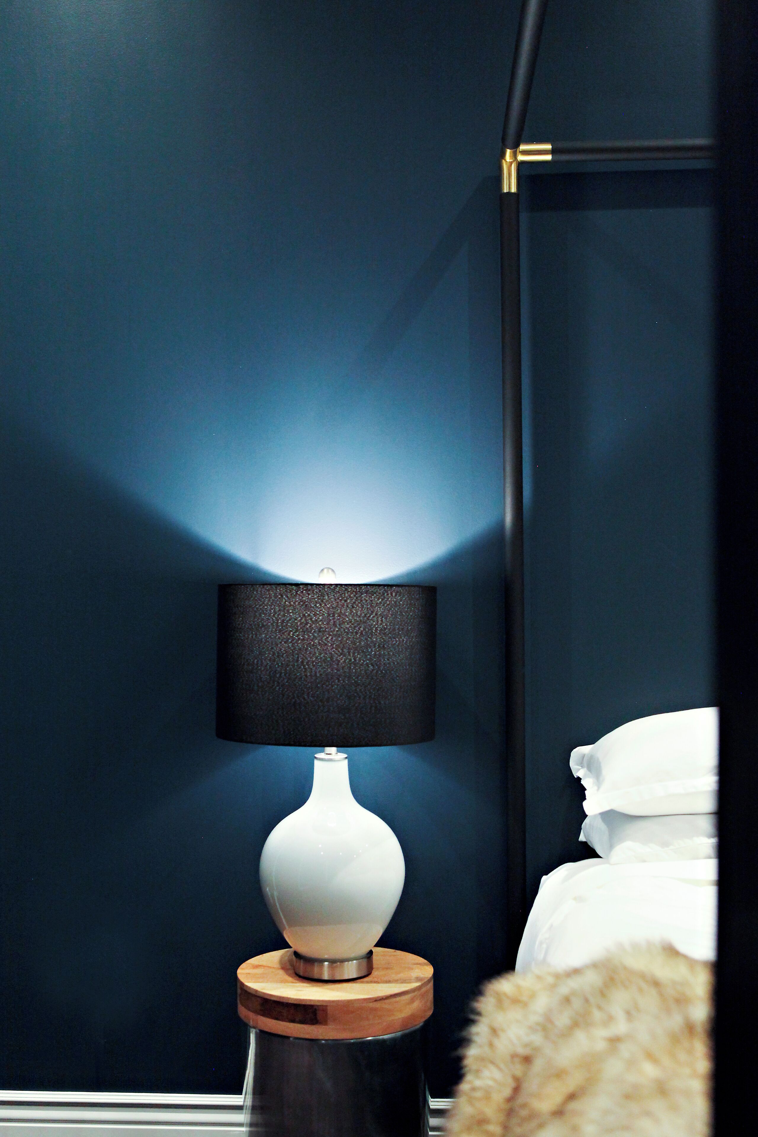 A bedside table with a white vase and a black lamp against a dark blue wall.