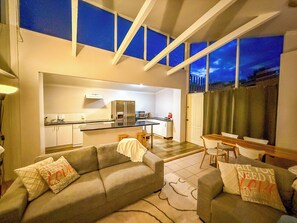An open-plan living and kitchen area with a sofa, a dining table, and a view of the city at dusk. - Cozy, well appointed unit near centre of Jindabyne and 30mins to snowfields. (Jindabyne)