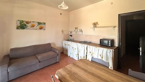 Een knus woongedeelte met een bank, een eettafel, een kitchenette met fornuis en broodrooster, en een schilderij aan de wand.
