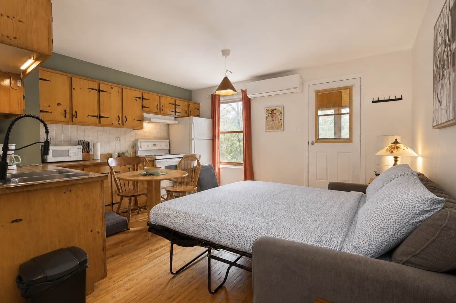 A compact room with a bed, a small dining table, wooden cabinets, a kitchenette, and a window.