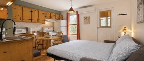 A compact room with a bed, a small dining table, wooden cabinets, a kitchenette, and a window.