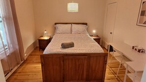 Un dormitori amb un llit de fusta, dues taules de nit amb làmpades, una làmpada penjant i un escriptori blanc amb una cadira.
