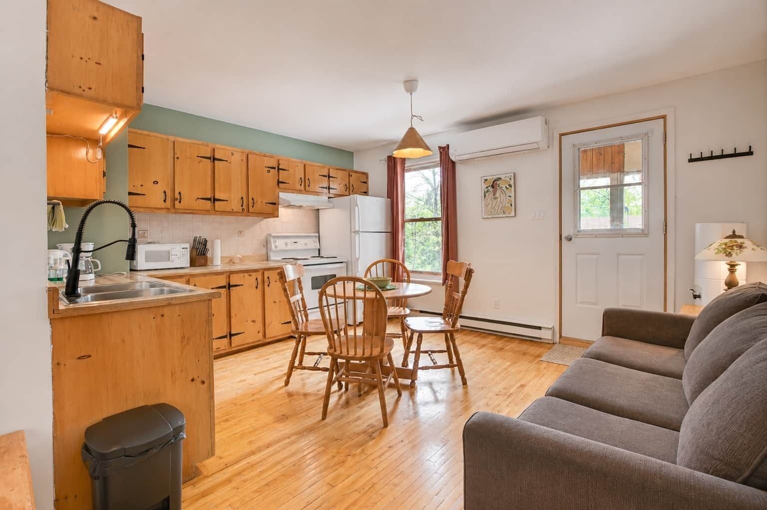 A compact kitchen with wooden cabinets, a dining table with chairs, a sofa, and a trash bin.