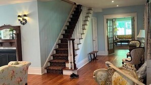 Un salón amplio con una escalera de madera, sillones estampados con motivos florales y una alfombra de diseño rojo.