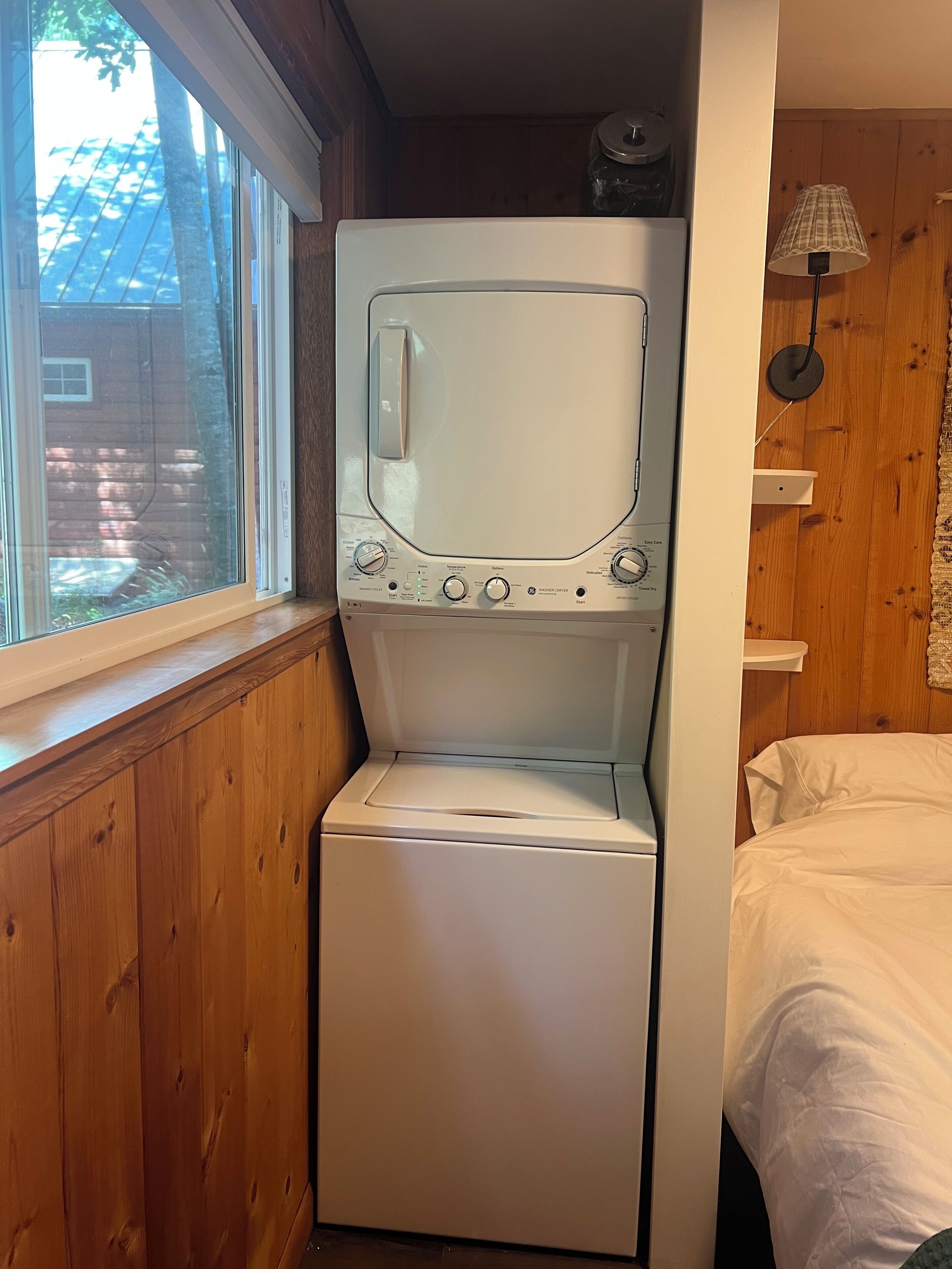 A compact washer and dryer unit in a small room with wooden walls, a window, and a lamp.