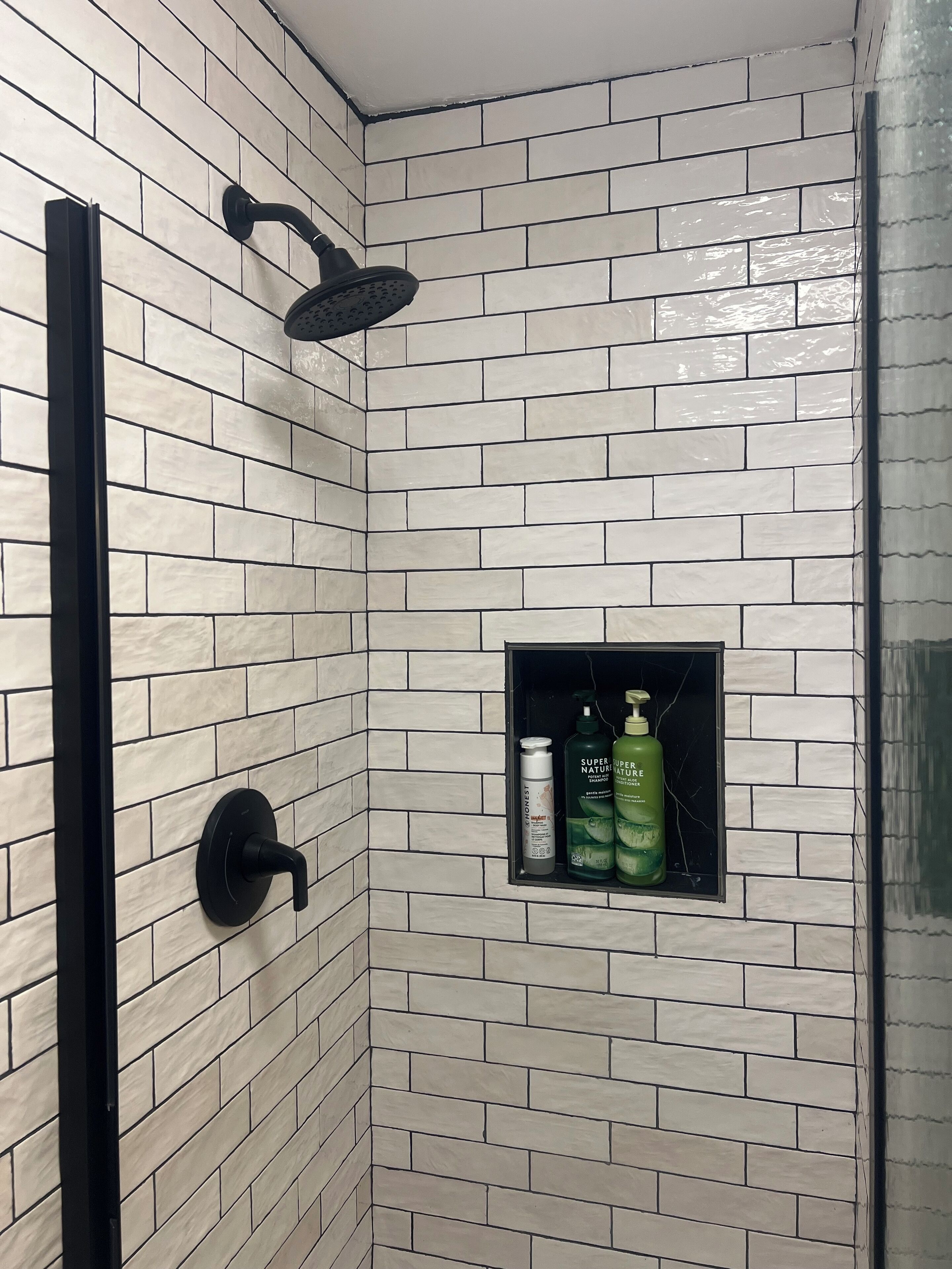 Ein Badezimmer mit weiß gefliesten Wänden, einem schwarzen Duschkopf und einem Regal mit zwei Shampoo- und Conditionerflaschen.