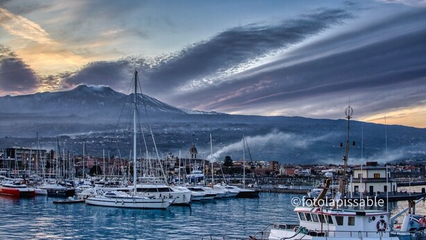 A marina with boats docked, a mountain in the background, and a cloudy sky. - Bedda Sicily (Riposto)