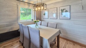 Un comedor con una mesa de madera, sillas y un jarrón con flores. Hay cuadros en la pared y un ventanal con vista a follaje.