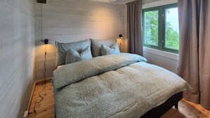 Un dormitorio con cabecera de madera, una cama grande con ropa de cama gris, dos lámparas de noche y una ventana con cortinas.