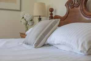A neatly made bed with white linens and two pillows, a wooden headboard, a bedside table with a lamp, and a vase with white flowers. - Juniper House -  a house that sleeps 17 guests  in 8 bedrooms (Kingsbridge)