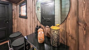 Una habitación con paredes de madera, una puerta negra, un espejo redondo, una mesa con un florero y un taburete.