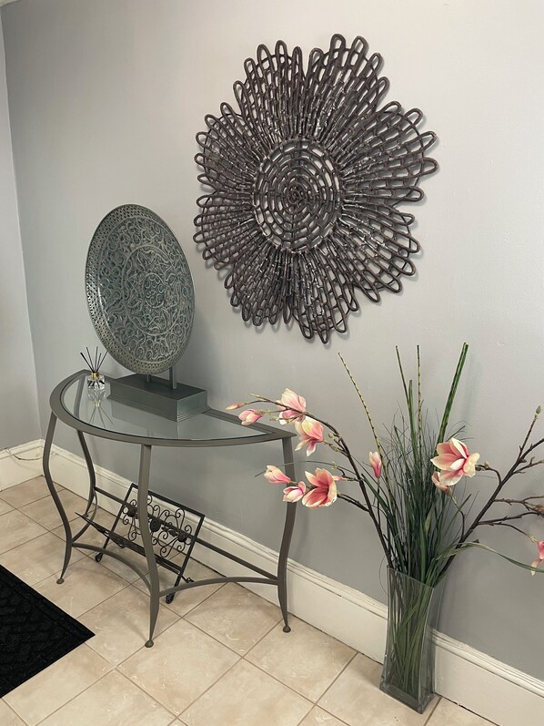 Una habitación con una mesa de vidrio, un adorno metálico en la pared, un florero con flores rosadas y un plato estampado sobre la mesa.