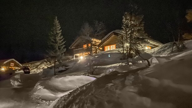 Eine verschneite Landschaft mit einer Hütte und einem größeren Gebäude mit beleuchteten Fenstern.