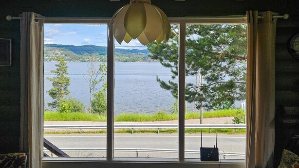 A room with a large window offering a view of a lake, a road, and greenery. - Amazing home in Svelvik with WiFi (Svelvik)