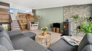 A modern living room with grey sofas, a dining area with a table and chairs, a stone fireplace, and a staircase leading to another level.