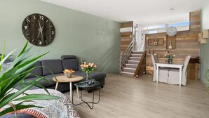 A modern living room with a large round clock, a grey sofa, a small round table with a bowl of fruit, and a staircase leading to a dining area.