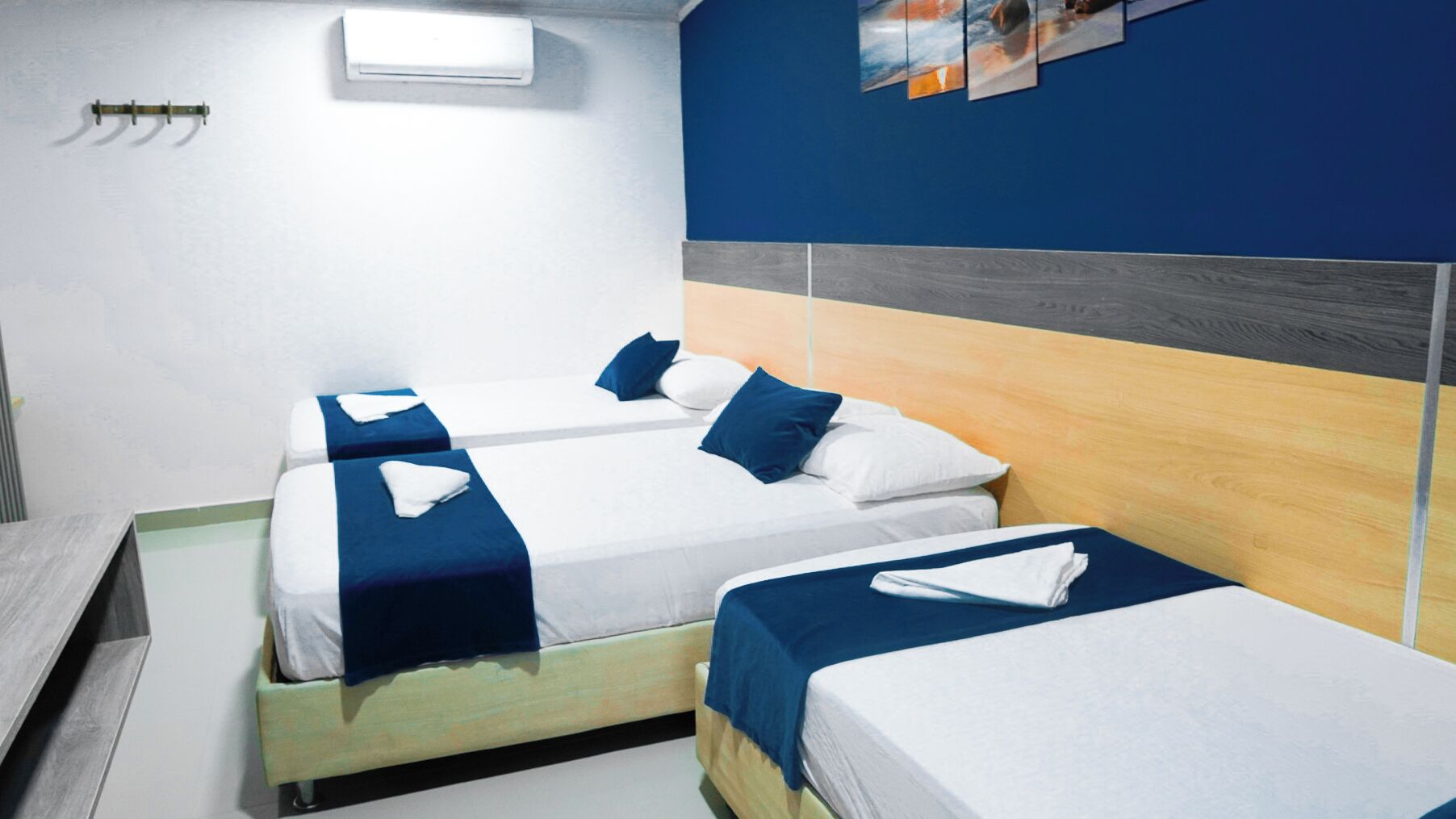Hotellihuone, jossa on kaksi erillistä sänkyä. Jokaisessa sängyssä on sininen peitto ja valkoiset tyynyt, puinen pääty sekä pieni valkoinen ilmastointilaite seinällä.