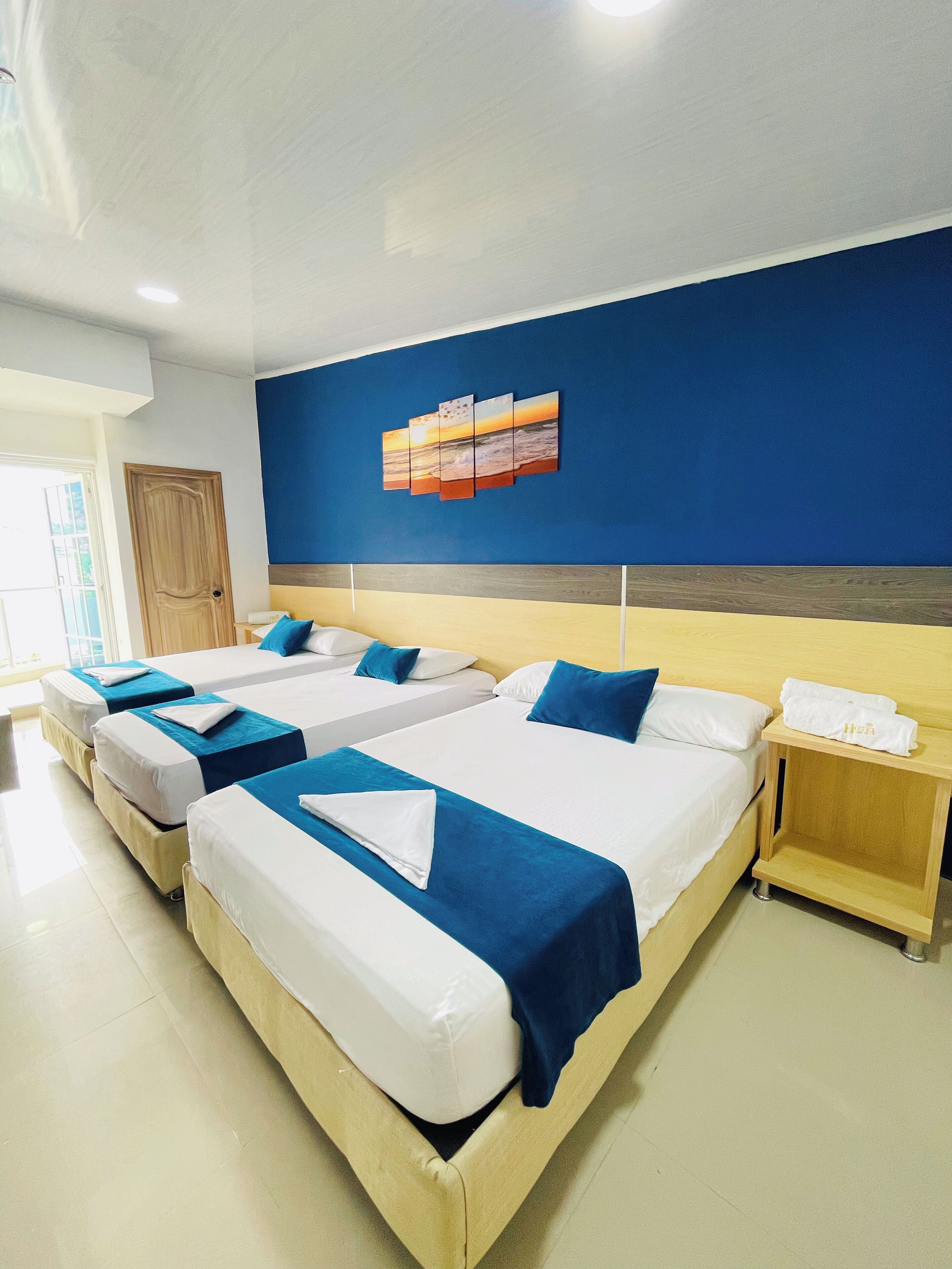 Ein Hotelzimmer mit drei Betten, jeweils mit weißer Bettwäsche und blauen Akzenten, einem hölzernen Kopfteil und einem Wandbild, das einen Sonnenuntergang über dem Meer zeigt.
