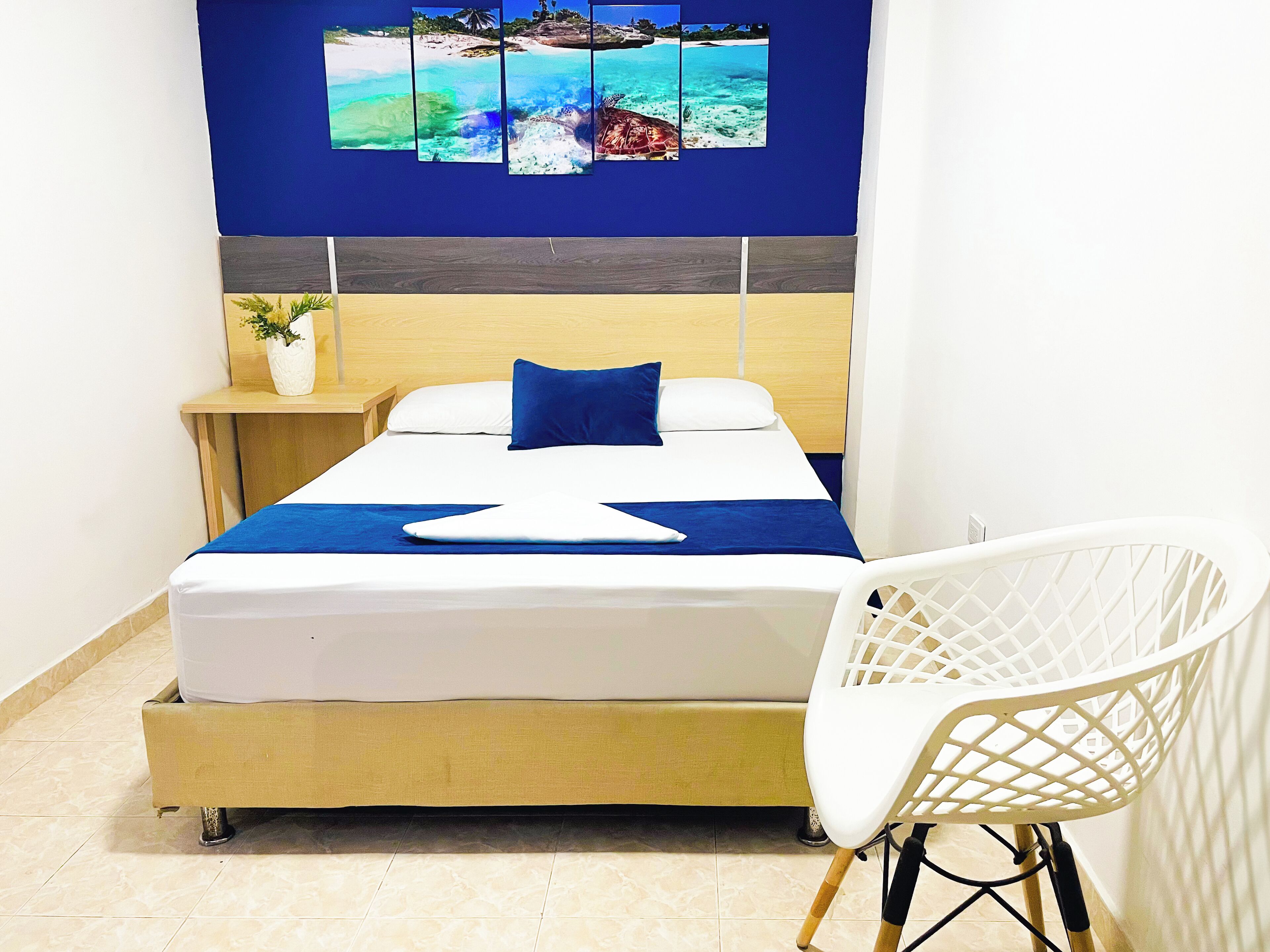 Een bed met een blauwe spreideken en kussens, een witte stoel, een nachtkastje met een plant en een muur met een schilderij van een tropisch strand.