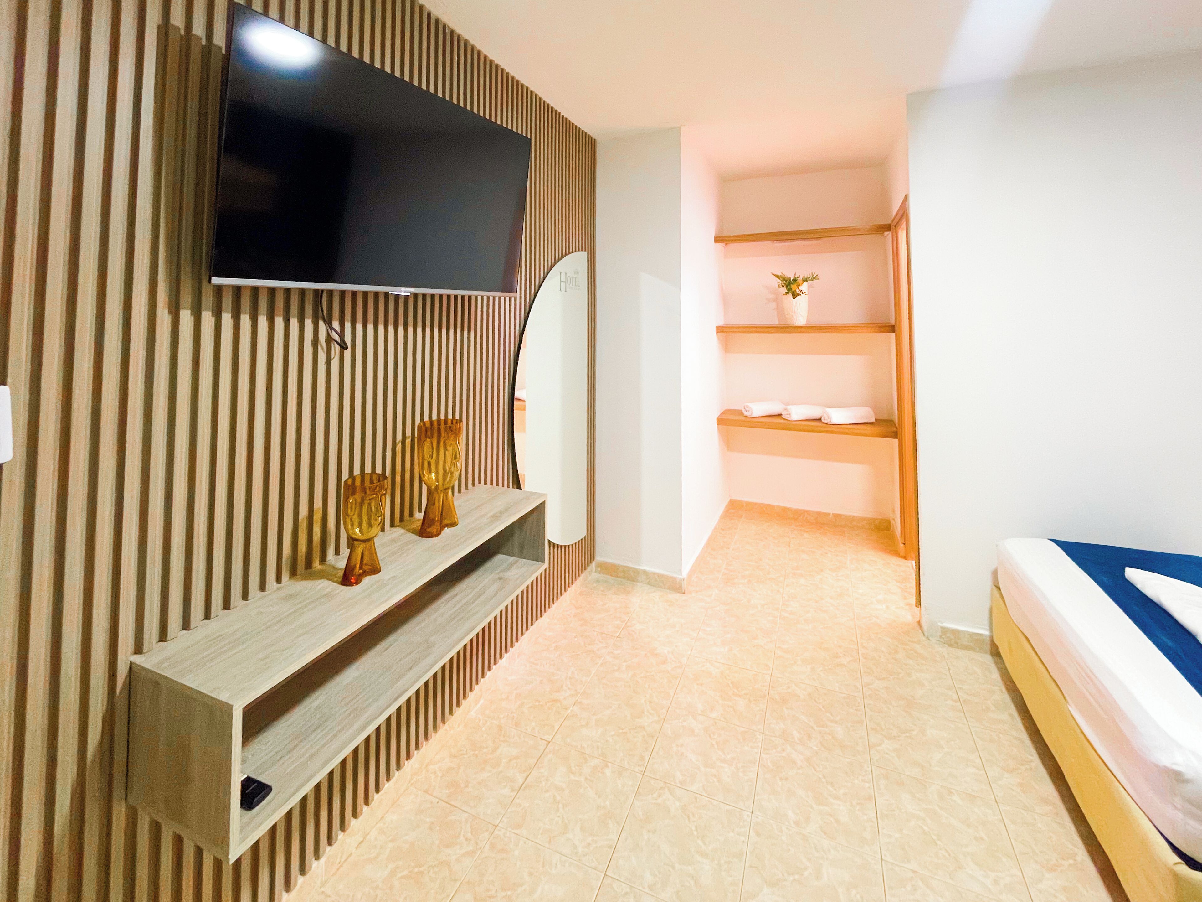 Habitación de hotel moderna con una televisión de pantalla plana montada en una pared de paneles de madera, una mesa consola de madera con objetos decorativos, una cama con colchón azul y un estante pequeño con toallas.