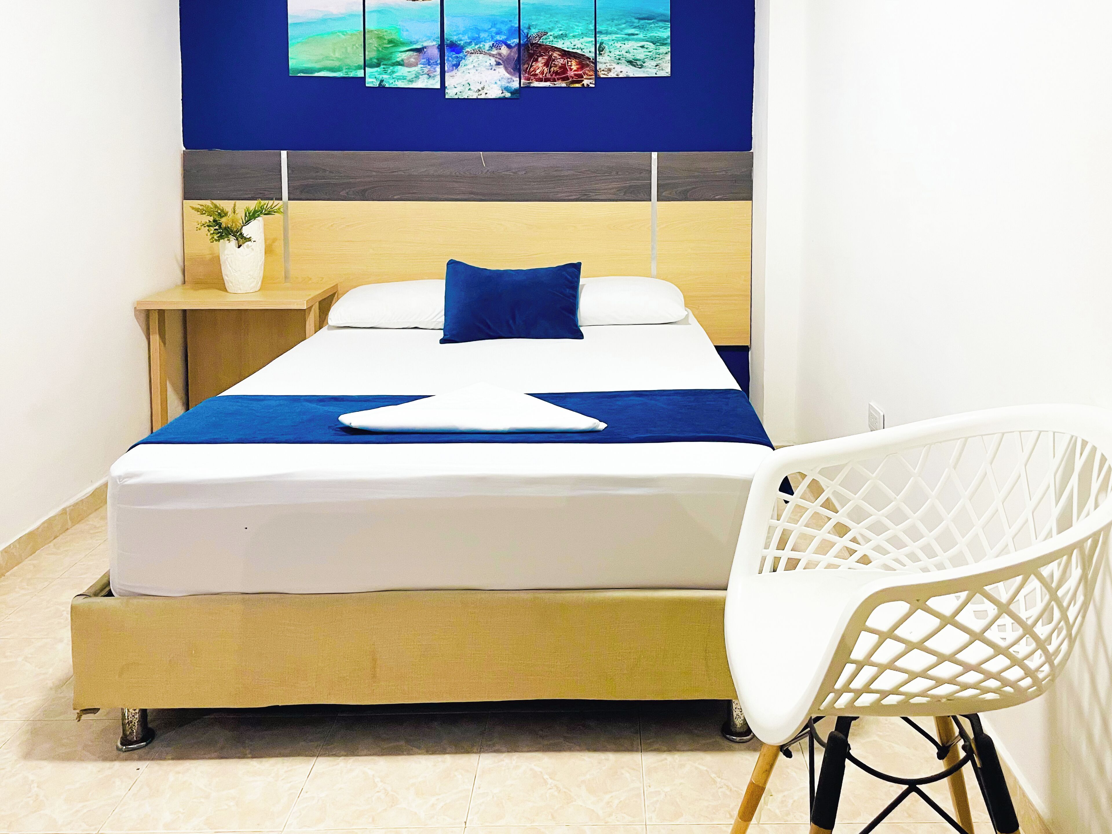 Un lit bien fait, recouvert d’une couvre-lit bleue et agrémenté d’oreillers, une chaise blanche au motif géométrique et une petite table d’appoint sur laquelle est posée une plante.