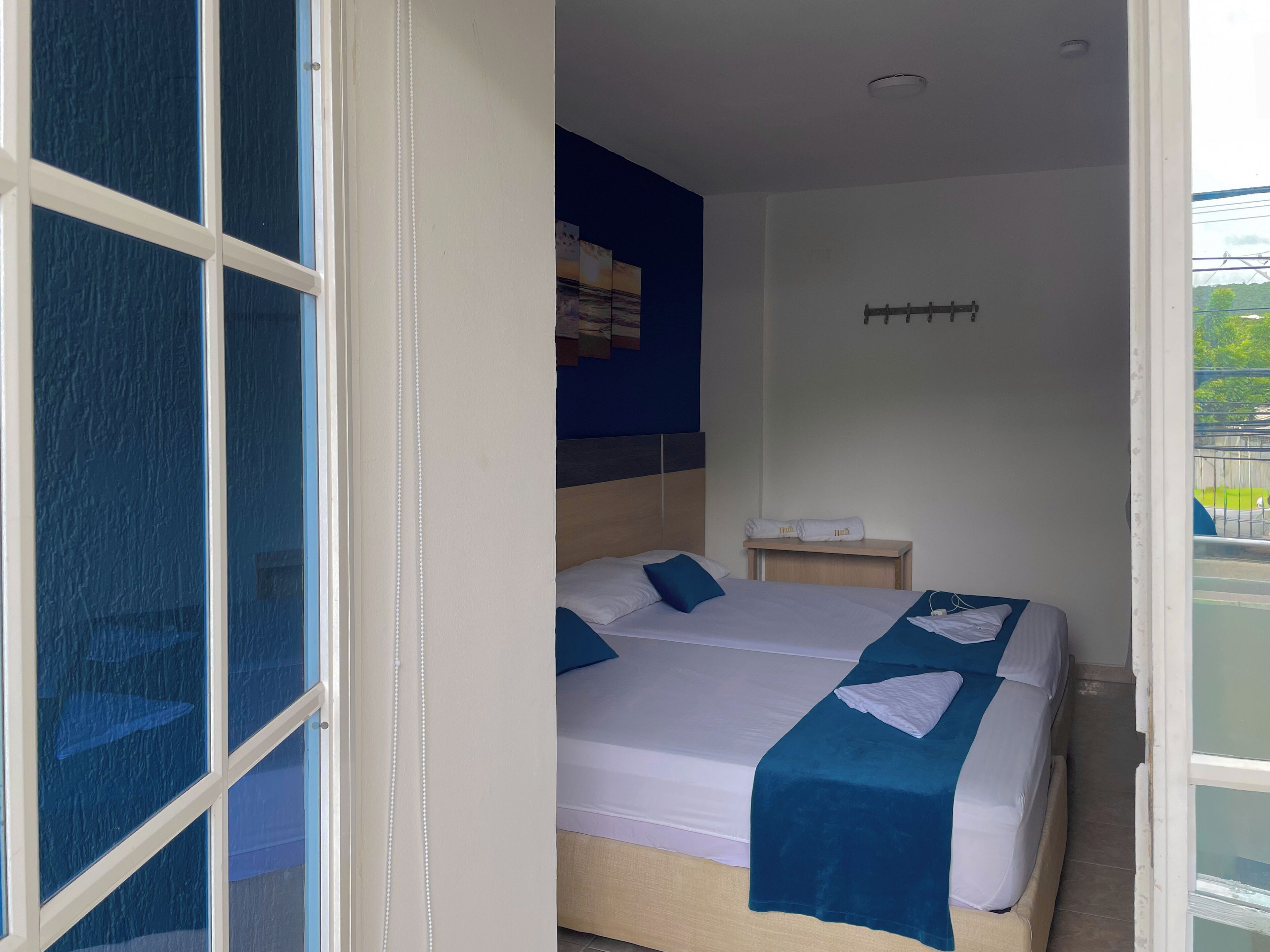 A hotel room with two beds, a small bedside table, a wall-mounted shelf, and a window with a view of the outdoors.