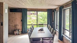 Uma sala de jantar com uma mesa comprida e cadeiras, janelas amplas com cortinas e vista para o verde lá fora.