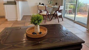 A modern living room with a dark wooden coffee table, a white vase with green plants, a dining area with a glass table and chairs, and a large window with a view of the outdoors. - Cozy Beachfront Getaway! (Los Angeles)