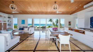 Amplia sala de estar con techo de madera, muebles blancos y un gran ventanal con vista al océano.