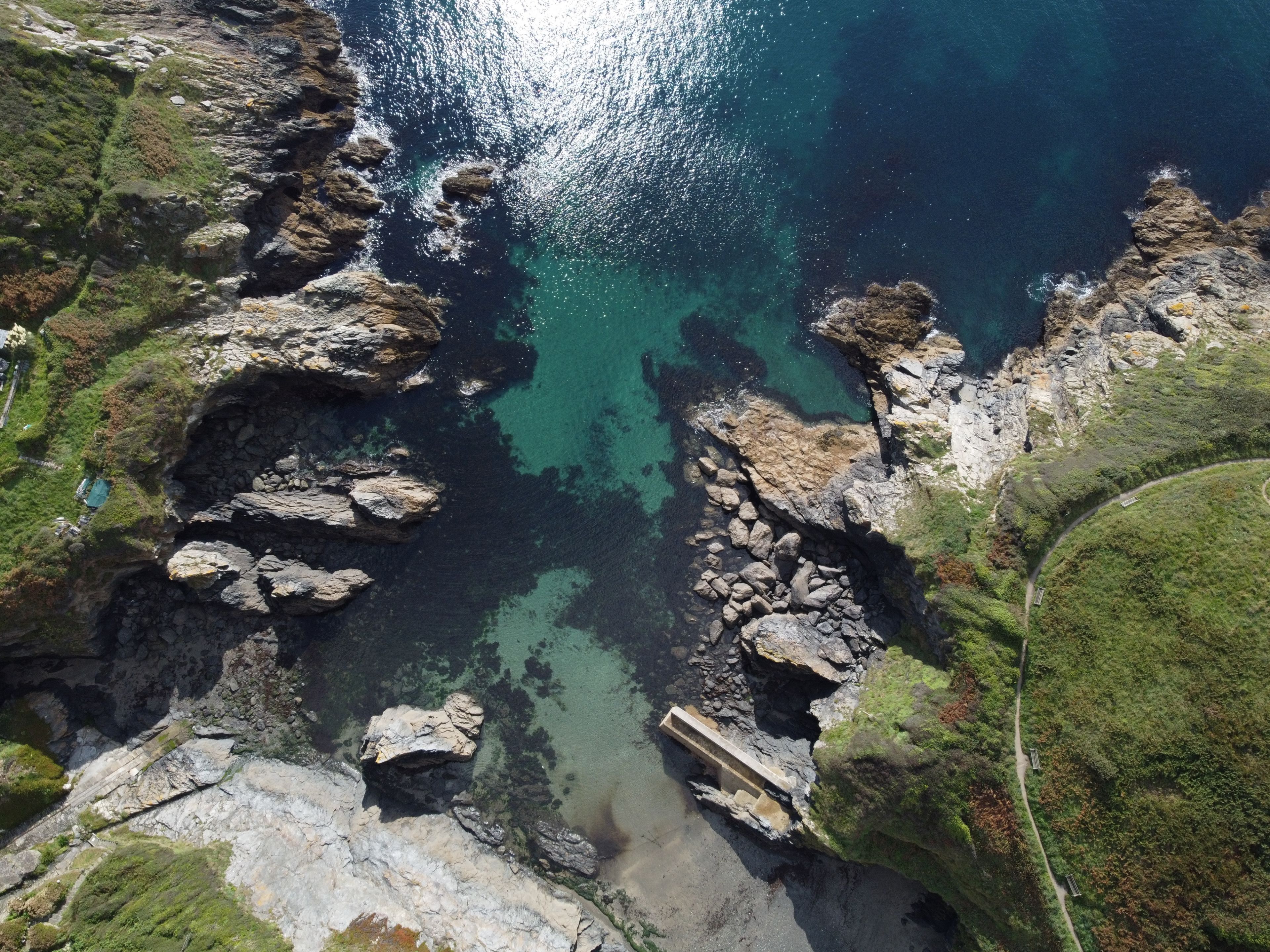 An aerial view of a coastal area with rocky terrain, clear water, and a paved path.