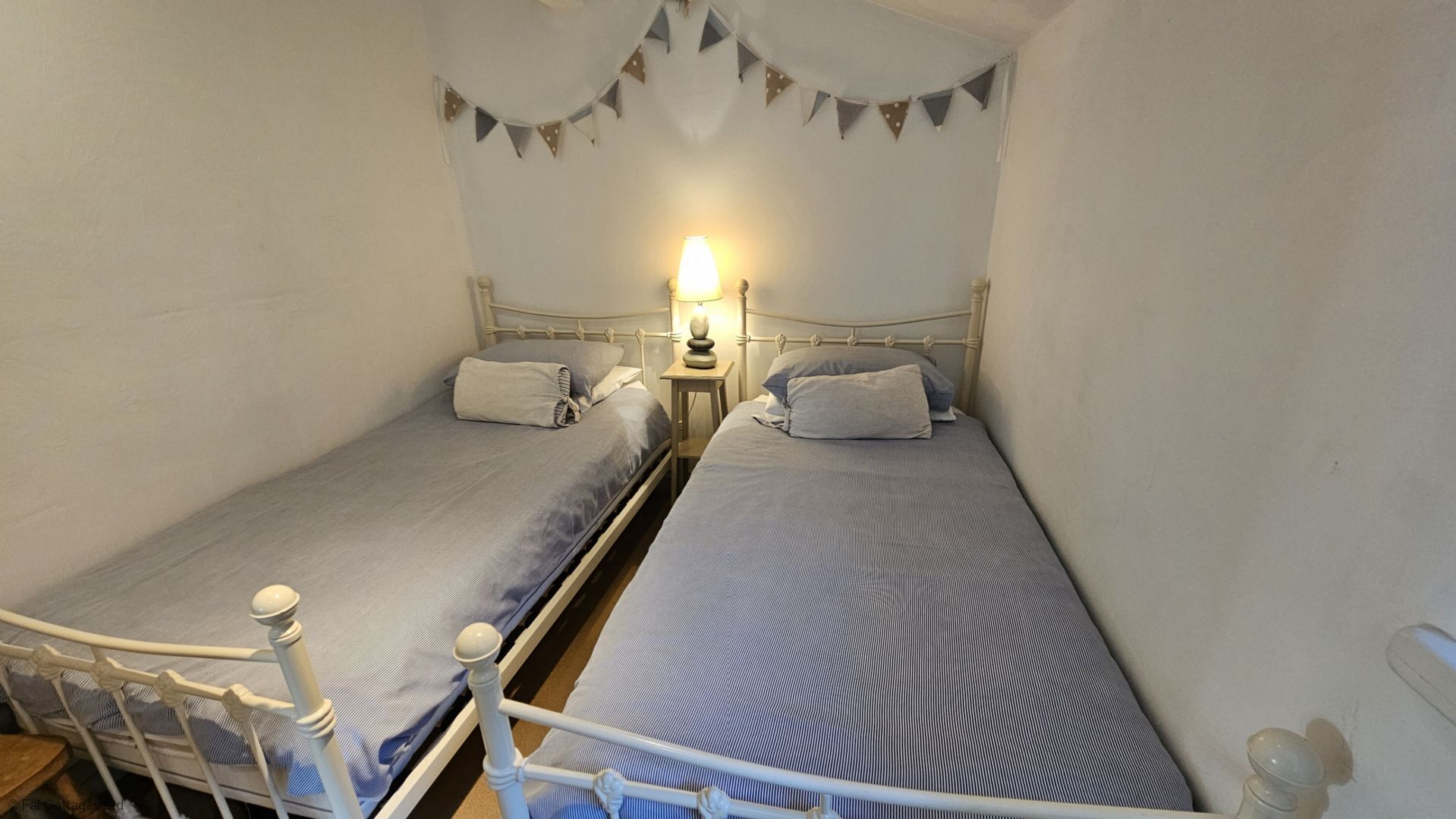 Un petit logement composé d’une seule pièce, comprenant deux lits simples, une table de chevet avec une lampe et une étagère fixée au mur.