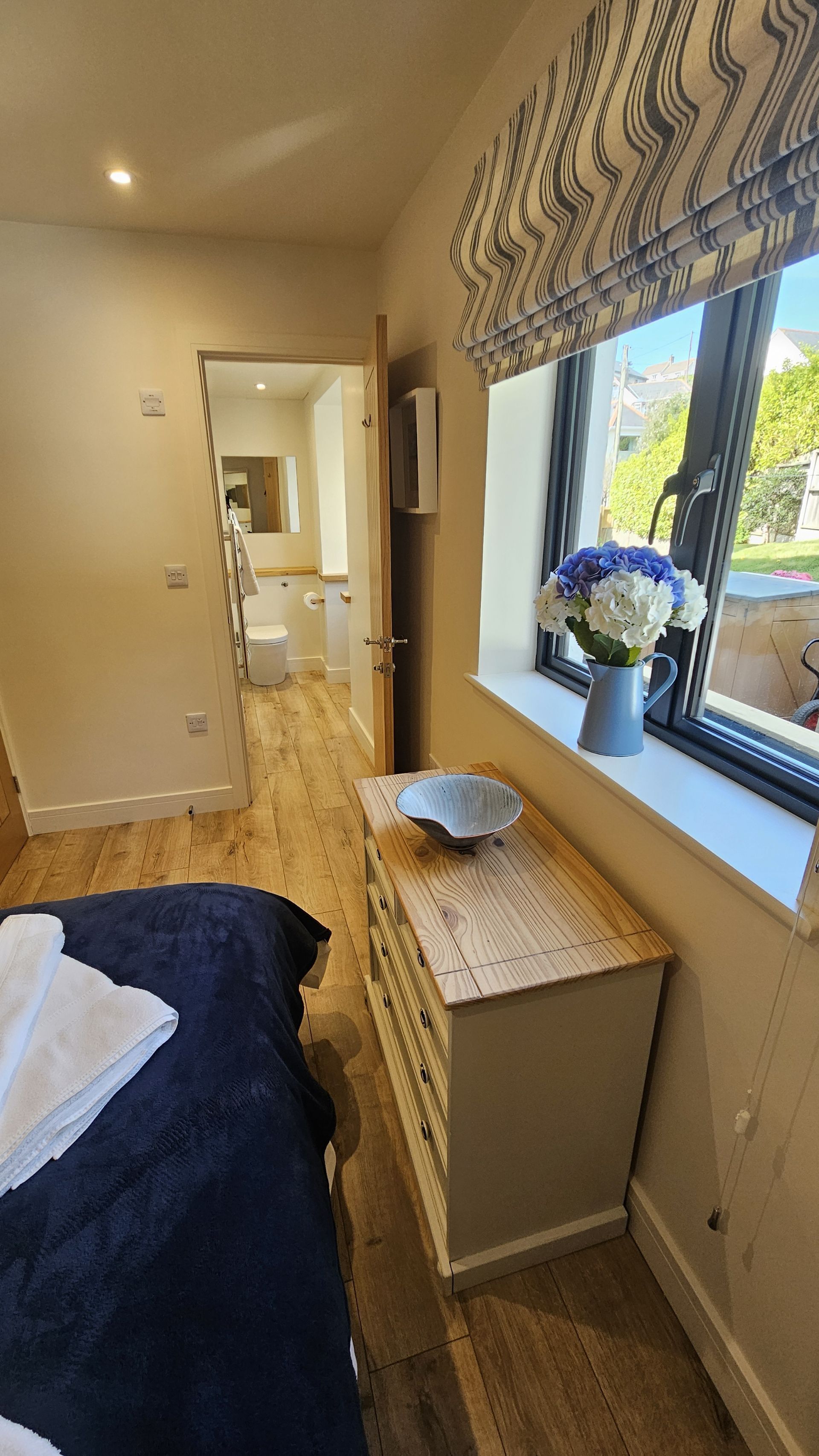 A bedroom with a wooden bed, a bedside table with a vase of flowers, a window with a view of the outdoors, and a door leading to a bathroom.
