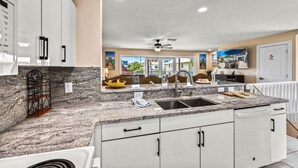 A modern kitchen with granite countertops, white cabinets, a double sink, and a ceiling fan. - Dolphin Resort | Private Pool, Spa, Tiki Bar, Game Room & 3 Min Walk to Beach (Holmes Beach)