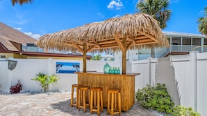 A thatched outdoor bar area with stools, a stone-paved patio, and a white fence. - Dolphin Resort | Private Pool, Spa, Tiki Bar, Game Room & 3 Min Walk to Beach (Holmes Beach)