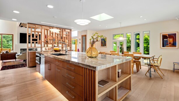Une cuisine moderne avec des meubles en bois, un plan de travail en marbre et un îlot central. On y trouve un coin repas avec des chaises et une table en bois, ainsi qu’une grande fenêtre qui laisse entrer la lumière naturelle.