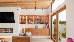 Un salon moderne avec un canapé en bois, une cheminée, un téléviseur fixé au mur et une grande fenêtre donnant sur l’extérieur.
