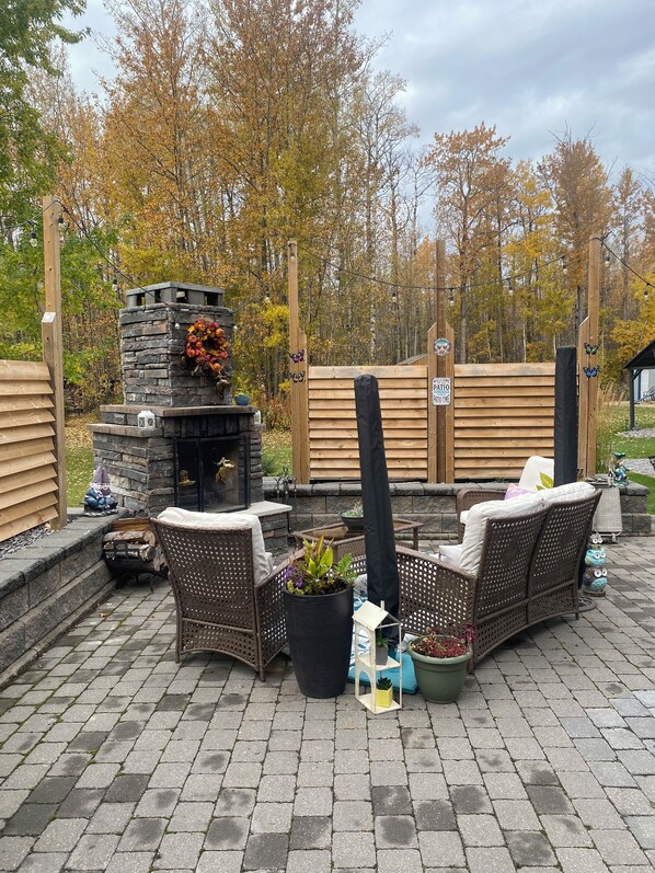 En udendørs terrasse med pejs af sten, fletmøbler og plantekasser.