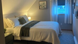 Un dormitorio con cama, mesita de noche, lámpara de pared, ventana con cortinas e imagen enmarcada en la pared.