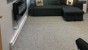 Una sala de estar con un sofá modular, un televisor de pantalla plana, dos ventanas con persianas y una chimenea.