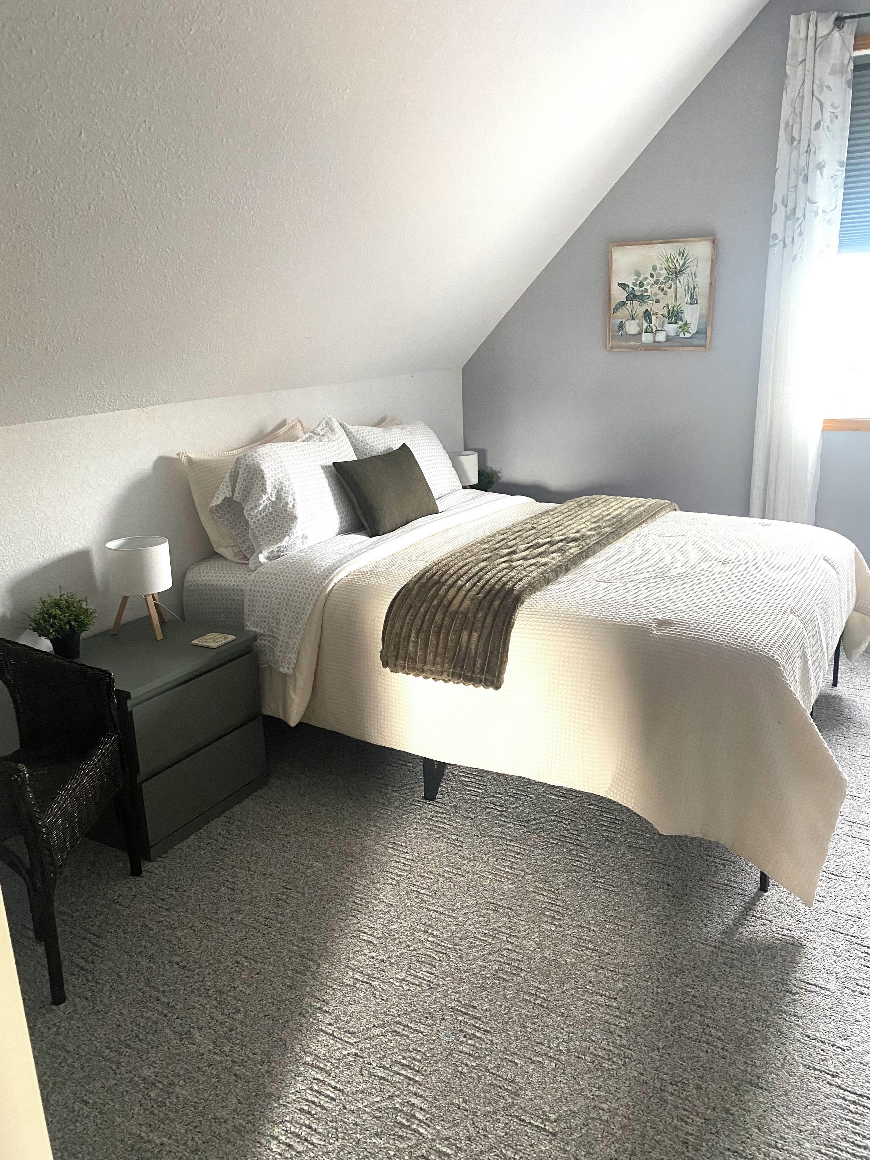 Un dormitorio con cama, mesitas de noche, lámpara, silla y una planta en la pared.
