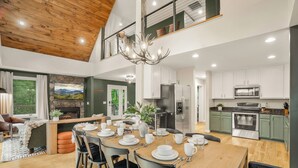 Une vaste salle à manger avec une table en bois dressée pour quatre personnes, entourée de chaises noires. On aperçoit une cheminée au fond de la pièce, et celle-ci est dotée d’un plafond haut avec des poutres apparentes.