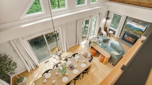 Un vaste espace de vie aménagé dans un style loft, avec une table à manger pour six personnes, un coin salon confortable doté d’une cheminée et de grandes fenêtres offrant une vue sur l’extérieur.