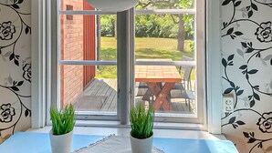 Ein Zimmer mit Fenster und Blick auf eine Terrasse mit Holztisch und -stühlen. Zwei Blumentöpfe stehen auf einer blauen Tischdecke.
